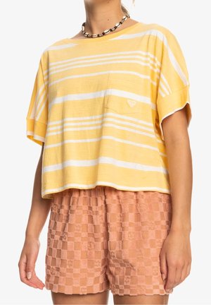 Frau trägt ein lockeres gelb-weiß gestreiftes Kurzarmhemd mit Brusttasche und strukturierte pfirsichkarierte Shorts.