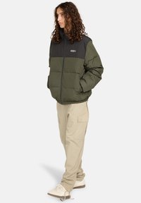 Giovane con capelli ricci che indossa un giubbotto imbottito nero e verde oliva, pantaloni cargo beige e sneakers bianche, in piedi con le mani in tasca.