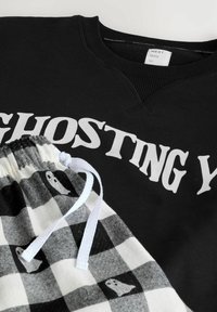Černá mikina s nápisem "GHOSTING YOU", spárovaná s kostkovanými kalhotami s motivy duchů a bílým šňůrkovým pásem v pase.
