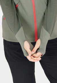 Olivgrüne Zip-Up-Jacke aus strukturiertem Stoff, mit rotem Reißverschluss und Daumenlöchern an den Ärmelbündchen. Trägt sie über schwarzen Hosen.