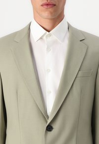 Chaqueta de traje verde claro con solapas de muesca, cierre con un solo botón negro y diseño sin costuras; combinada con una camisa blanca de cuello por debajo.