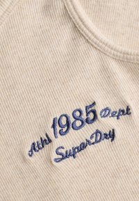 Tejido de canalé beige con texto bordado en azul marino "Auth 1985 Dept SuperDry" cerca de un escote redondo.