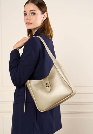 Jeune femme portant un blazer bleu marine tient un sac à bandoulière en cuir beige avec un fermoir doré, debout devant un mur clair.