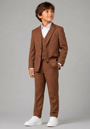 Garçon portant un costume marron avec un gilet, une chemise blanche et des baskets blanches, debout avec une main dans la poche, regardant sur le côté.