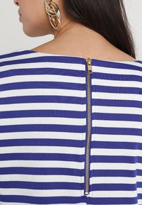 Gestreepte top met blauwe en witte horizontale lijnen, met een gouden rits aan de achterkant en een brede halslijn voor gemakkelijk aan- en uittrekken.