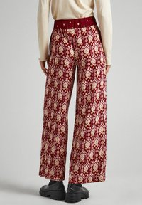 Breda byxor i burgunderfärgad tyg, med ett intrikat mönster i kräm- och gult, med resårmidja och svarta klumpiga stövlar.