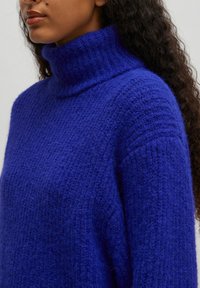Royal blue turtleneck sweater made of soft, textured knit fabric, featuring a relaxed fit and wide ribbed collar.