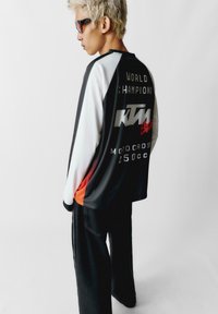 Čierno-biely dlhý rukáv dres s textom "SVETOVÍ ŠAMPIONI", logom KTM a tučným písmom "MOTOCROSS 250cc". Model má na sebe tmavé nohavice.