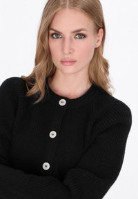 Schwarze Strickjacke mit geripptem Design, runder Halsauschnitt, Knopfverschluss mit hellen Knöpfen und langen Ärmeln.