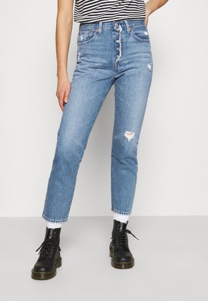 Femme portant un jean déchiré taille haute bleu clair avec braguette boutonnée, chaussettes blanches, bottines noires lacées et un haut rayé noir et blanc.
