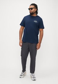 Μπλε ναυτικό μπλουζάκι με γραφικό σχέδιο, σε συνδυασμό με γκρι παντελόνι cargo και λευκά αθλητικά παπούτσια. Το μοντέλο φοράει γυαλιά ηλίου και έχει πλεξούδες στα μαλλιά.