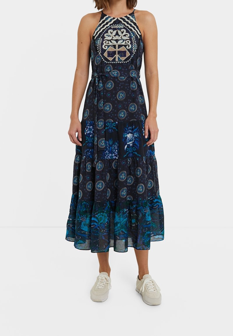 Femme portant une robe midi sans manches, à motifs bleu marine avec des dessins floraux et géométriques, et des baskets blanches sur un fond blanc.