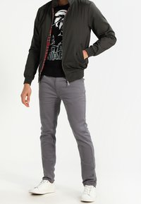 Szara kurtka typu bomber z prążkowanymi mankietami, noszona na czarnej koszulce z nadrukiem. Szare, dopasowane spodnie i białe trampki dopełniają strój.