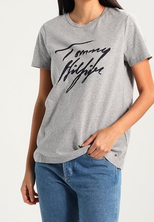 Harmaa puuvillainen t-paita, jossa on musta, kursivoitu "Tommy Hilfiger" -logo. Lyhyet hihat ja pyöreä kaulus. Yhdistetty sinisiin farkkuihin.