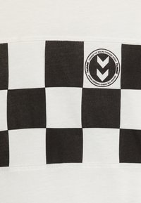 Hvid T-shirt med et sort og hvidt ternet mønster hen over brystet, med et rundt logo og tekst på højre side.