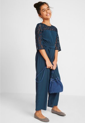 Ragazzina sorridente che indossa una tuta blu navy con maniche in pizzo, tiene una borsa a secchiello blu coordinata e indossa ballerine grigie.