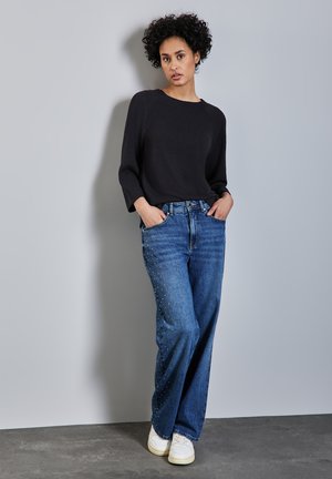 Schwarzer Strickpullover mit lockeren Ärmeln, kombiniert mit blauen Jeans aus Denim mit Nietenverzierung und weißen Sneakers vor einem grauen Hintergrund.