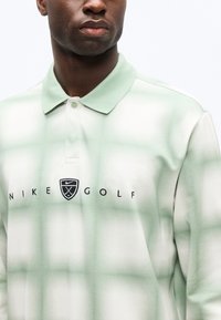 Άνδρας που φοράει μπλουζάκι πόλο Nike Golf σε ανοιχτό πράσινο και λευκό καρό, με γιακά και λωρίδα με κουμπιά, το λογότυπο και το κείμενο είναι κεντραρισμένα στο στήθος.