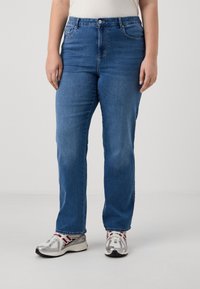 Persona vestida con vaqueros de pierna recta azules, una camiseta sin mangas blanca y zapatillas deportivas blancas con detalles en rojo y plateado, de pie sobre un fondo neutro.