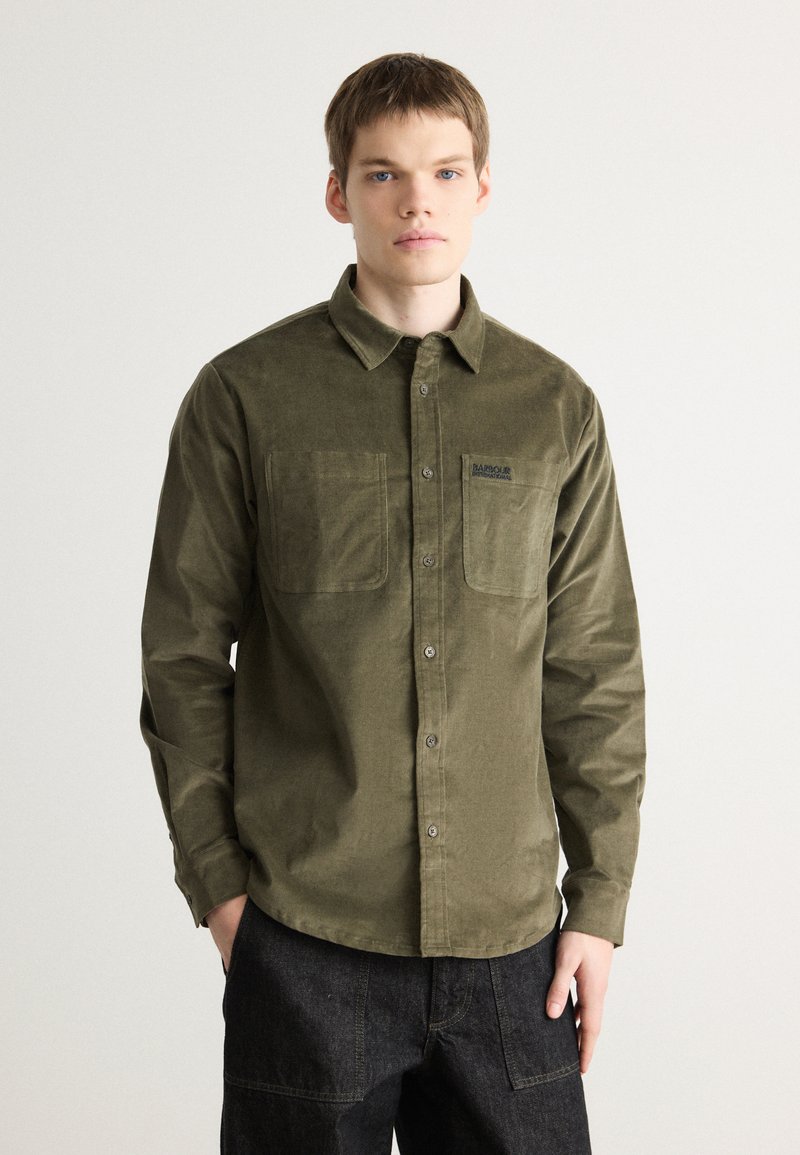 Olive green corduroy shirt with a button-down collar, two chest pockets, and a logo on the pocket. Textured fabric with a relaxed fit.