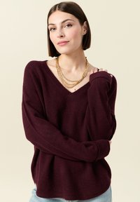 Burgundy v-ringad tröja i mjukt, texturerat tyg med överdimensionerade ärmar, i kombination med lager av gyllene halsband. Ljus bakgrund framhäver detaljer.