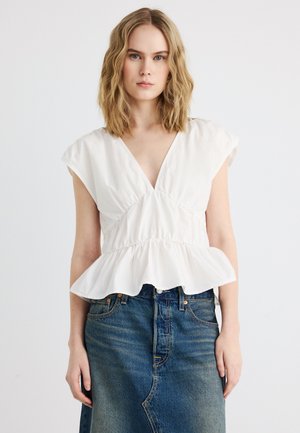 Femme aux cheveux blonds ondulés portant un haut péplum blanc à col en V et une jupe en denim bleu délavé sur un fond clair uni.