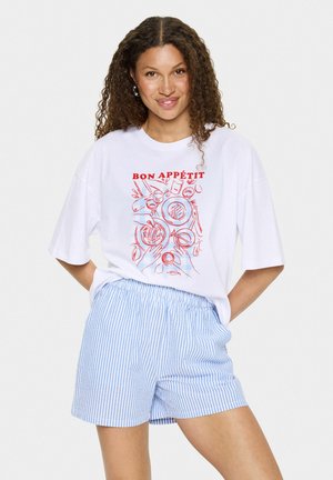 T-shirt blanc avec le texte "BON APPÉTIT" en rouge et des illustrations de nourriture. Associé à un short rayé bleu clair et blanc. Style décontracté, coupe relax.