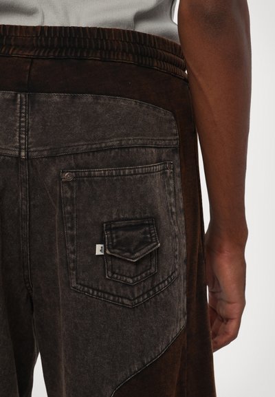 Bruine spijkerbroek met een gestructureerd uiterlijk, voorzien van een achterzak met een decoratieve klep en zichtbare stikseldetails. Inclusief elastische tailleband.