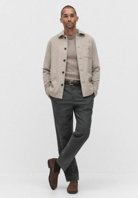 Beige ullblandad jacka med knäppning och två framfickor, i kombination med en ljusbrun tröja och mörkgrå byxor. Bruna mockaskor.