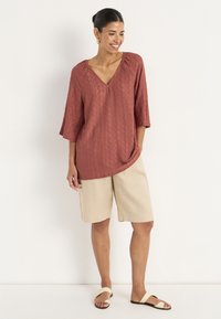 Rustrød, tekstureret bluse med V-hals og trekvartærmer, parret med beige linnedshorts og tan farvede slidesandaler.