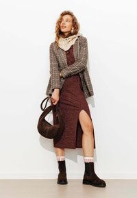 Blazer à carreaux marron sur une robe bordeaux avec une fente sur le côté. Bottines en suède marron avec des chaussettes blanches à motifs. Sac hobo marron. Fond clair.