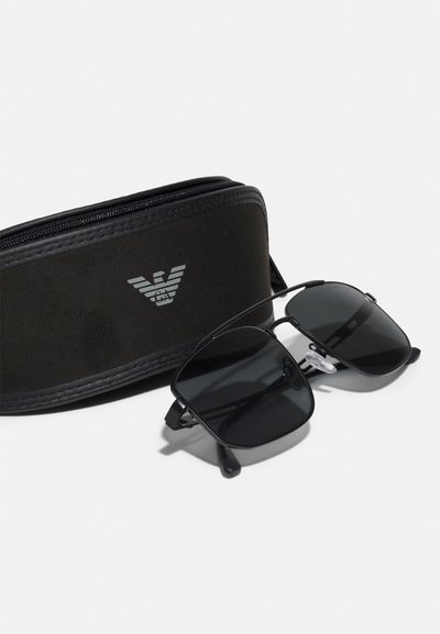 Occhiali da sole con lenti nere, montatura quadrata e cuscinetti nasali in gomma, insieme a una custodia nera con un logo. Texture liscia, chiusura a zip.