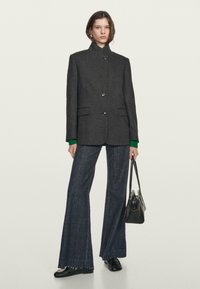 Szary oversize żakiet z wełny z kołnierzem stojącym oraz dwoma przednimi kieszeniami, w połączeniu z szerokimi, ciemnoniebieskimi dżinsami i czarną torebką.