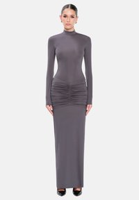 Mujer de pie con un vestido gris largo y ajustado con detalles fruncidos en las caderas, mangas largas y tacones negros sobre un fondo blanco.
