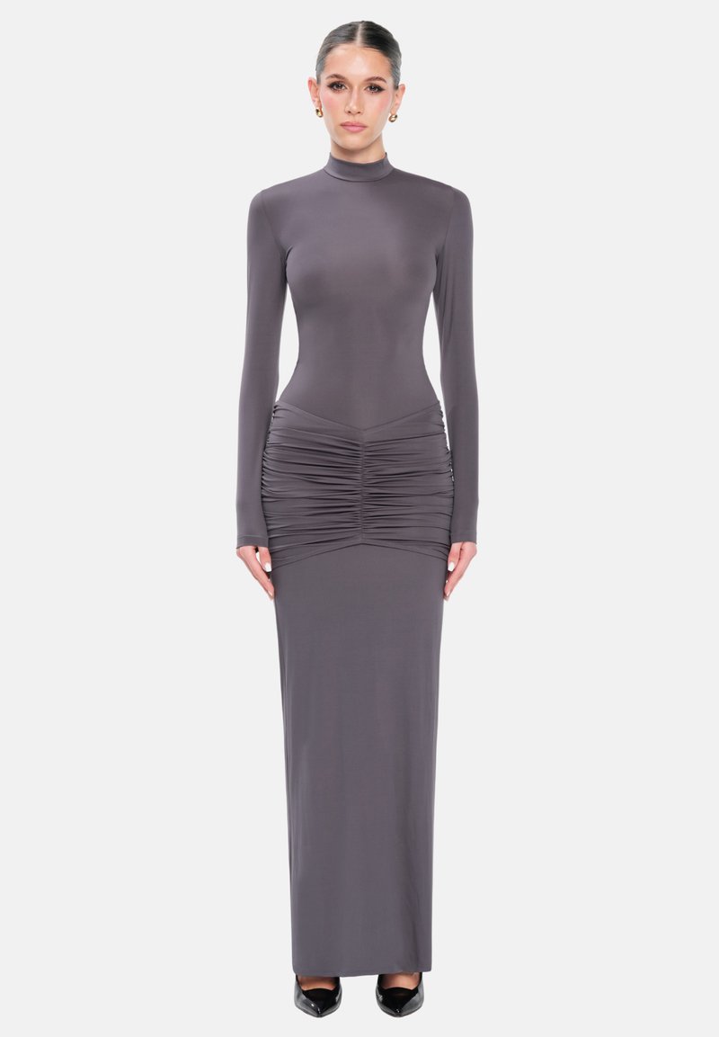 Mujer de pie con un vestido gris largo y ajustado con detalles fruncidos en las caderas, mangas largas y tacones negros sobre un fondo blanco.