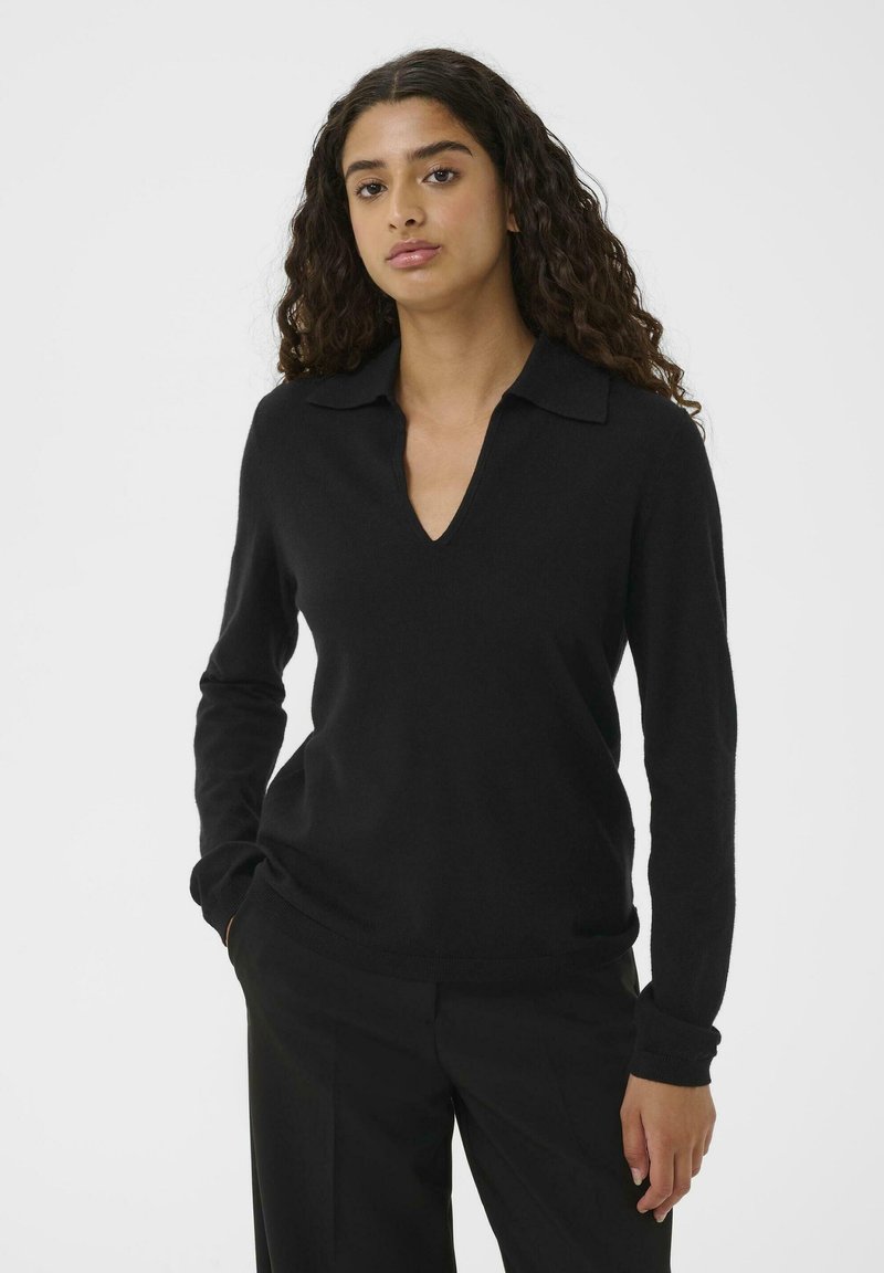 Donna con lunghi capelli ricci, che indossa un maglione nero a maniche lunghe con scollo a V e pantaloni neri, in piedi con una mano in tasca.