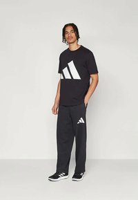 Black t-shirt with large white Adidas logo, paired with black sweatpants featuring smaller logo. Black sports shoes complete the outfit.