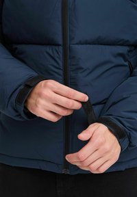 Mørkeblå dunjakke med lynlås foran, der har sorte velcro-manchetter. Stoffet ser glat og isoleret ud med quiltede sektioner.