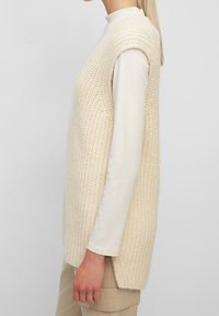 Pull sans manches en maille crème, avec un motif texturé, des fentes sur les côtés et une coupe ample, porté avec une chemise à manches longues en dessous.