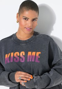 Sudadera gris de gran tamaño con la frase "BÉSAME" impresa en un degradado de naranja a púrpura. Textura suave, puños acanalados y un corte relajado.