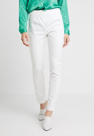 Femme portant un pantalon blanc slim, des chaussures blanches à talons et un chemisier vert à manches longues avec des pois noirs, debout devant un fond uni.