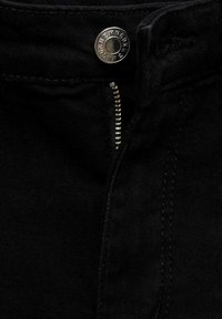 Gros plan sur un jean en denim noir avec une fermeture éclair argentée partiellement ouverte et un bouton argenté portant le nom de la marque.