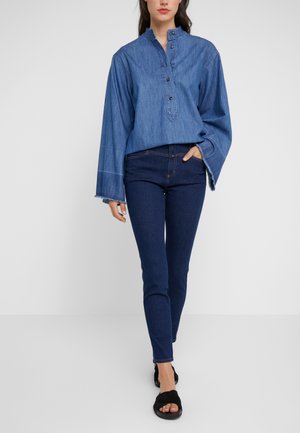 Kobieta ubrana w luźną niebieską jeansową bluzkę z szerokimi rękawami, ciemne obcisłe dżinsy i czarne sandały wsuwane z odkrytymi palcami, stoi z jedną ręką w kieszeni.