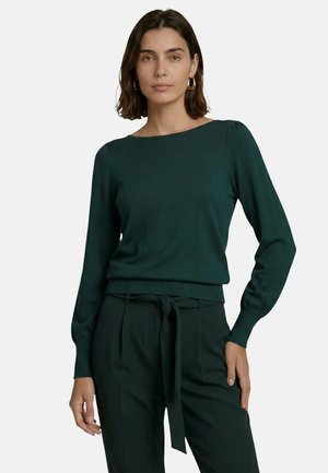 Femme aux cheveux châtains mi-longs porte un haut à manches longues vert foncé et un pantalon taille haute avec une ceinture en tissu assortie, debout devant un fond uni.