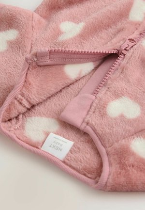 Rosa baby overall med mjuk fleece-textur. Har vita hjärt-mönster, en dragkedja och en tygetikett i nederkant.