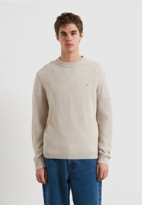 Beiger Strickpullover mit strukturiertem Muster, Rundhalsausschnitt und gerippten Bündchen, ausgestattet mit einem kleinen orangefarbenen Logo auf der linken Brust. Kombiniert mit blauer Denim.