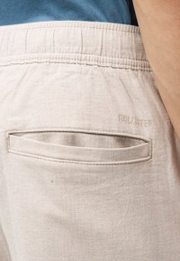 Κοντινό πλάνο σε τσέπη μπεζ παντελονιού με ελαστική μέση και διακριτικό λογότυπο Holister, φορεμένο με μπλε πουκάμισο μέσα και εμφανές χέρι.