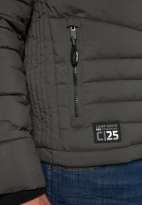 Chaqueta acolchada gris con costuras en forma de cuadrícula horizontal, un bolsillo con cremallera y un parche negro con la marca "CAMP DAVID" y "C25".