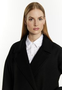 Zwarte wollen jas met een uitgesneden revers, gedragen over een witte blouse met kraag. Glide textuur en open voorkant ontwerp. Model met lang haar.