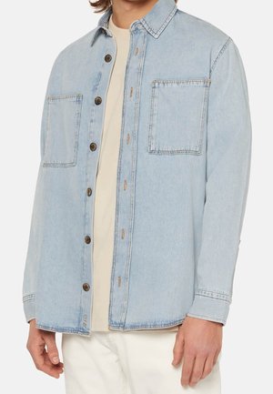 Chemise en denim bleu clair à boutons, avec deux poches poitrine, portée par-dessus un haut beige et un pantalon blanc.